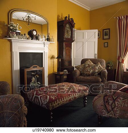 Stock Image - Longcase clock beside white fireplace in yellow livingroom with kelim upholstered stool . Fotosearch