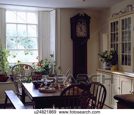 Stock Photo - Longcase clock beside white shutters at window in dining room with small table and Windsor chairs and cream dresser. Fotosearch