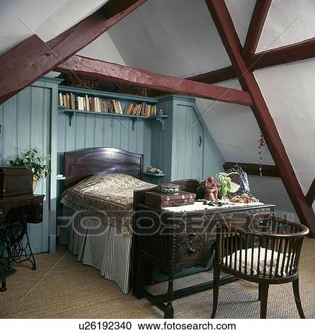 Stock Image - Mahogany bed below shelves in alcove on blue panelled wall in attic bedroom with small antique desk and chair . Fotosearch