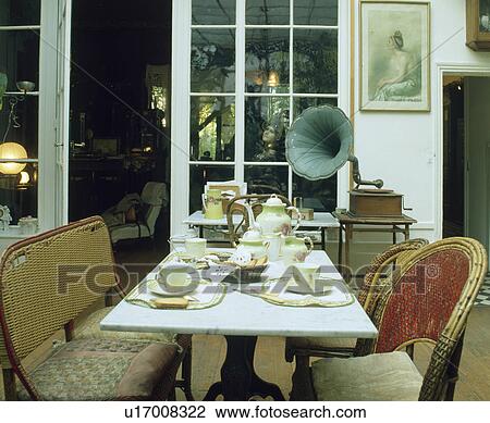 Stock Image - Marble top table set for tea in dining room with cane furniture and phonograph on table. Fotosearch