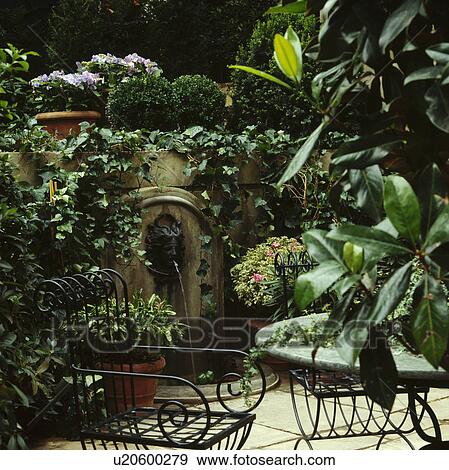 Stock Photo - Metal chairs and table on patio with wall fountain in country garden in summer. Fotosearch
