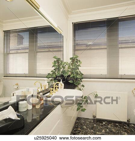 Stock Image - Mirror above white basin set into black-topped fitted white vanity unit in modern white bathroom with black slatted blind. Fotosearch