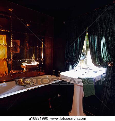 Stock Image - Mirrored wall above bath in black bathroom with dark green curtains above white pedestal basin. Fotosearch
