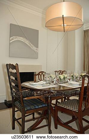 Stock Image - Modern circular light fitting above place settings on antique table with matching chairs in modern dining room. Fotosearch