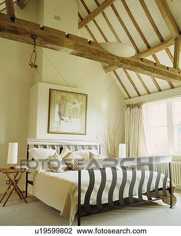 Stock Image - Modern metal bed with cream quilt and cushions in cream bedroom with beamed apex ceiling. Fotosearch
