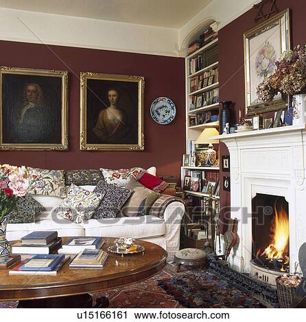 Stock Image - Oil paintings above cream sofa piled with cushions in brown living room with lighted fire in fireplace. Fotosearch