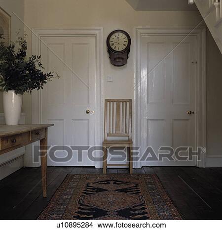 Picture - Old clock above wooden chair between white internal doors in country hall with oriental rug. Fotosearch