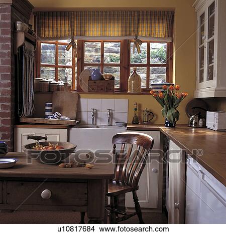 Picture - Old pine table and chairs in yellow cottage kitchen with checked blind at window above white Butler's sink. Fotosearch