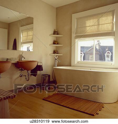 Stock Photo - Oval concrete bath and Philippe Starck basins in modern bathroom with cream linen blind and wooden mat on maple flooring. Fotosearch