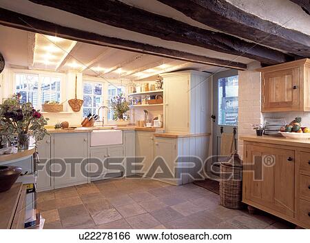 Stock Photograph - Pale grey fitted cupboards in cottage kitchen with beamed ceiling and quarry floor tiles. Fotosearch