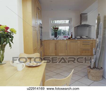 Pale wood table and chairs and fitted cupboards in modern kitchen dining room with tall cacti on white tiled floor View Large Photo Image Stock Photo - Pale wood table and chairs and fitted cupboards in modern kitchen dining room with tall cacti on white tiled floor. Fotosearch