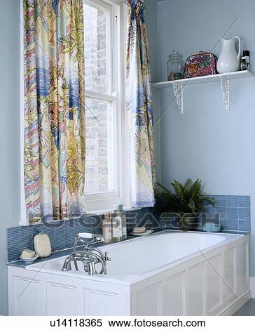 Stock Photography - Patterned curtains at window above white panelled bath in pastel blue bathroom. Fotosearch