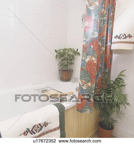 Stock Image - Patterned plastic curtain on bath with green houseplants in small bathroom. Fotosearch