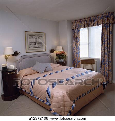 Peach Quilt With Blue White Striped Inserts On Bed With Upholstered Grey Headboard In Bedroom With Floral Curtains Stock Photograph