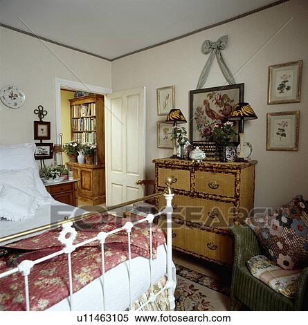 Stock Photography - Picture above lamps on bamboo chest-of-drawers in cottage bedroom with red floral quilt on antique brass bed. Fotosearch