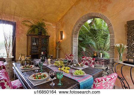 Picture - Pink chairs & colourful cloth on table with place settings on covered veranda with arched doorway. Fotosearch