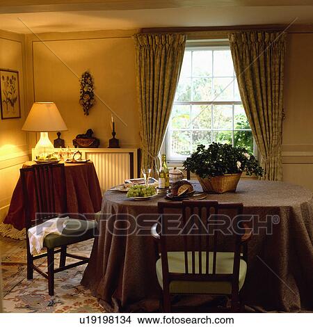 Place setting and houseplant on table with brown cloth in country dining room with lighted lamp on table in the corner View Large Photo Image Picture - Place setting and houseplant on table with brown cloth in country dining room with lighted lamp on table in the corner. Fotosearch