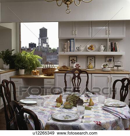 Stock Photo - Place settings on patchwork cloth on circular table in white kitchen with view of city through window. Fotosearch