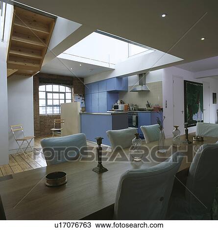 Stock Image - Rectangular dining table and upholstered chairs in openplan loft conversion with blue kitchen units and wooden flooring. Fotosearch