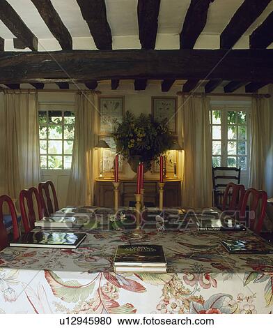 Stock Image - Red candles on floral tablecloth in country living room with beamed ceiling and lighted lamps between windows. Fotosearch