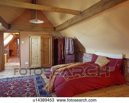Stock Image - Red duvet on bed in attic bedroom with patterend rug and pine wardrobe . Fotosearch
