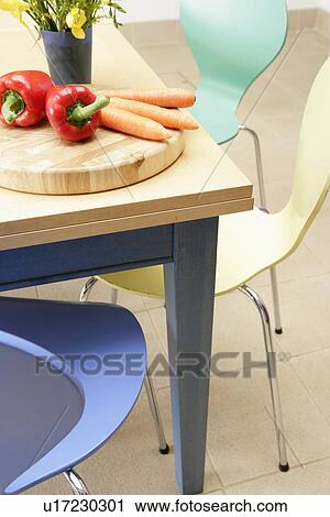 Stock Image - Red peppers and carrots on chopping board on modern wooden table with pastel Art Jacobsen-style chairs. Fotosearch