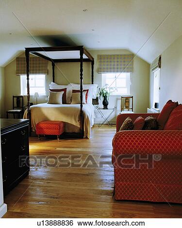 Stock Photograph - Red sofa in large white attic bedroom with simple four-poster bed and wooden flooring. Fotosearch
