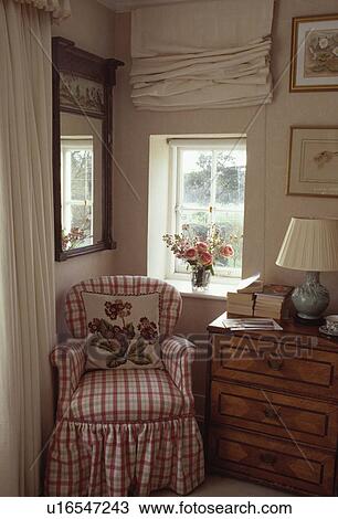 Stock Image - Red+white checked armchair and antique chest-of-drawers below small cottage window with cream blind. Fotosearch
