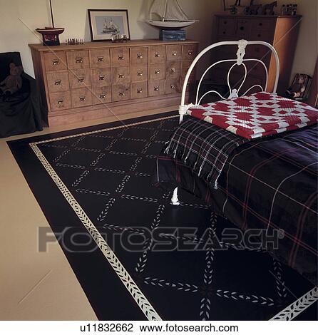 Stock Image - Red+white patchwork quilt and tartan rugs on single white wrought-iron bed in bedroom with painted rug on floor. Fotosearch