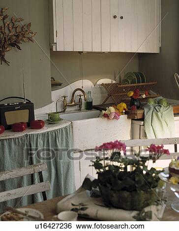 Stock Photograph - Roberts radio on worktop above curtained cupboard beside white ceramic sink in cottage kitchen. Fotosearch
