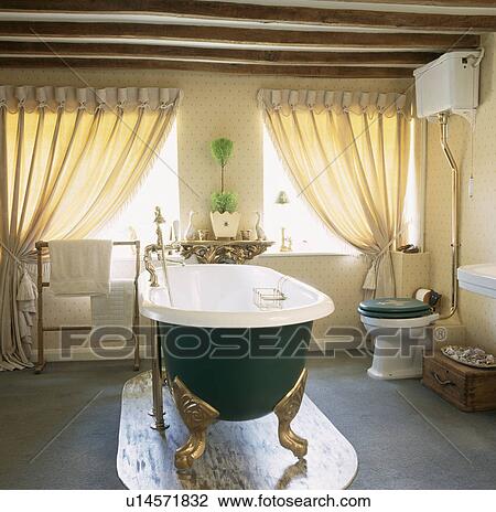 Stock Image - Rolltop bath with gold clawfeet on marble base in townhouse bathroom with cream curtains and wallpaper. Fotosearch