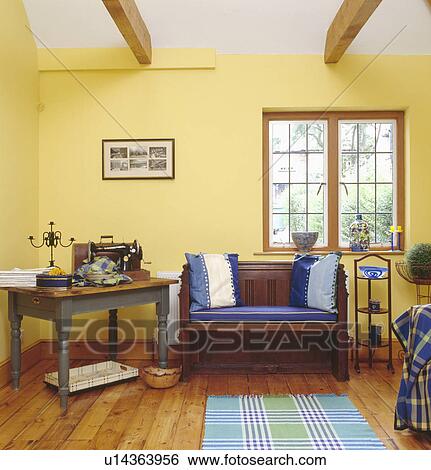 Sewing machine on table beside wooden settle with blue cushions below window in pastel yellow workroom View Large Photo Image Stock Photograph - Sewing machine on table beside wooden settle with blue cushions below window in pastel yellow workroom. Fotosearch