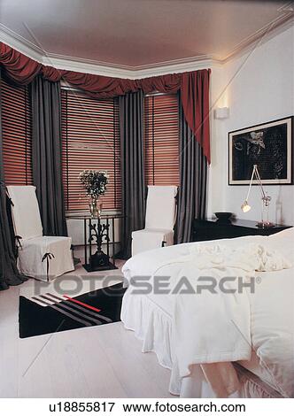 Slatted Wooden Blinds And Grey Curtains With Red Pelmets On Bay Window In Bedroom With White Chairs And Carpet Stock Photo