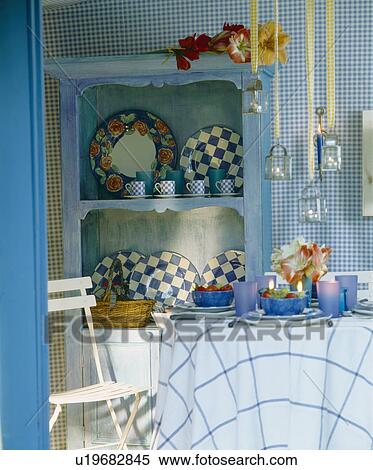 Stock Photography - Small circular table with checked cloth and blue glass and crockery in garden shed with checked plates on blue shelves. Fotosearch