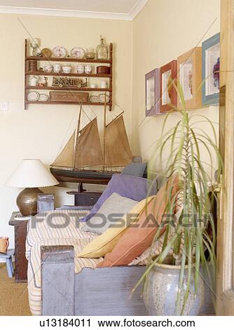 Stock Image - Small set of shelves above antique model boat in corner of living room with pastel cushions on the sofa. Fotosearch