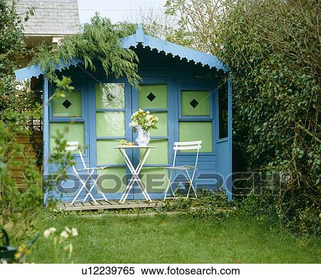 Stock Photography - Small white table and chairs in front of green and blue painted summerhouse. Fotosearch