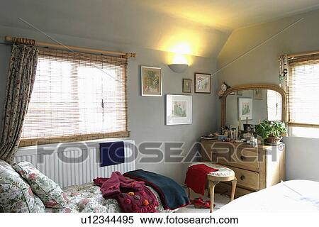 Stock Photography - Split cane blinds on window above radiator in small grey bedroom with mirror above pine chest-of-drawers. Fotosearch