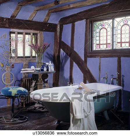 Stock Photograph - Stained glass window above turquoise freestanding rolltop bath in mauve bathroom with beamed walls. Fotosearch