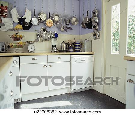 Stock Image - Stainless steel pans on wall above electrical appliances in mauve kitchen with fitted white cupboards. Fotosearch