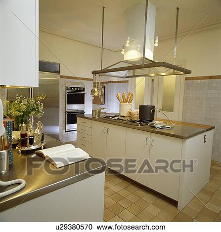 Stock Image - Stainless steel worktops in modern white kitchen with large extractor fan above rectangular island unit. Fotosearch