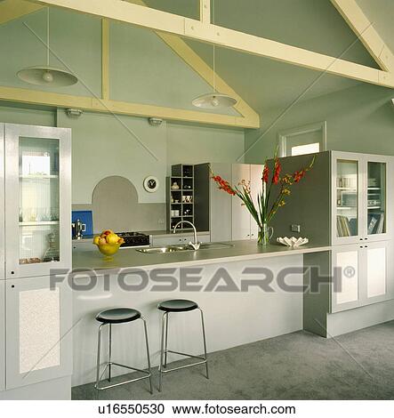 Stock Image - Stools at breakfast bar in modern grey-green kitchen with cream beams on apex ceiling. Fotosearch