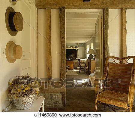 Stock Image - Straw hats on wall in beamed cottage hall with wicker chair beside doorway with view of living room. Fotosearch