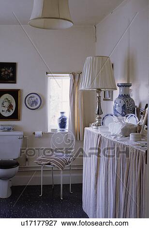 Striped cushion on metal chair in white cottage bathroom with lamp on vanity unit with blue+white striped curtains View Large Photo Image Stock Photo - Striped cushion on metal chair in white cottage bathroom with lamp on vanity unit with blue+white striped curtains. Fotosearch