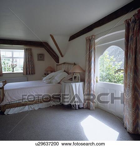 Stock Image - Sunny country bedroom with grey carpet and circular table with cloth beside bed with striped duvet. Fotosearch
