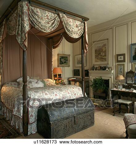 Stock Image - Swagged floral caonpy on four-poster bed in cream panelled country bedroom with studded antique chest . Fotosearch