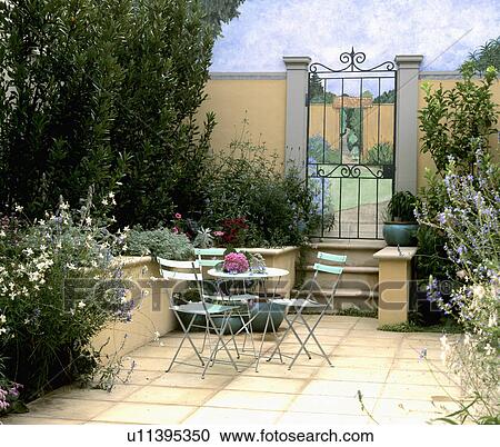 Stock Image - Table and pastel green chairs on patio in walled garden with summer-flowering shrubs and metal gate. Fotosearch