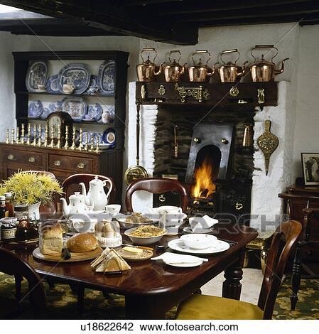 Stock Image - Table set for tea in cottage dining room with collection of copper kettles on shelf above fireplace. Fotosearch