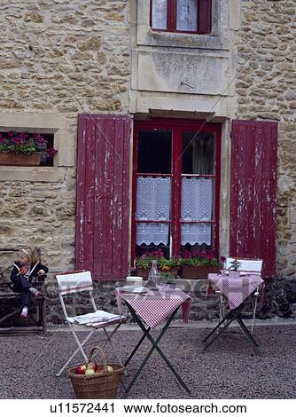 Stock Image - Tables and chairs in front of traditional country house in France with red shutters. Fotosearch