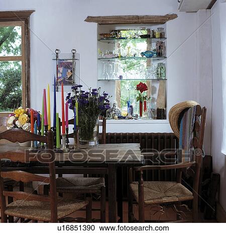 Stock Image - Tall colourful candles on rustic wooden table in white dining room with glass shelves across window. Fotosearch