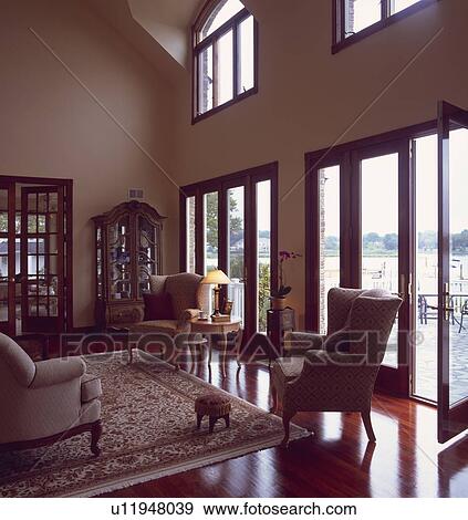 Stock Photo - Traditional armchairs and patterned rug on polished wood floor in double height livingroom with view through glass doors doors to waterside patio. Fotosearch